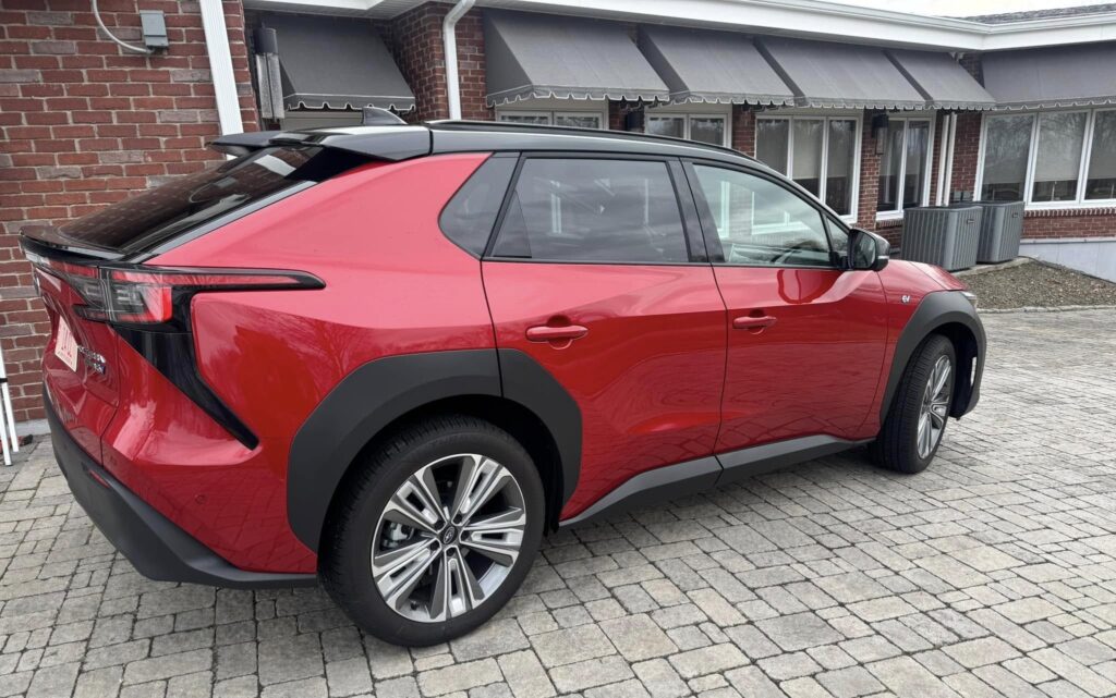 Sleek red 2025 Subaru Solterra SUV with black roof and trim, parked on patterned stone driveway near brick building
