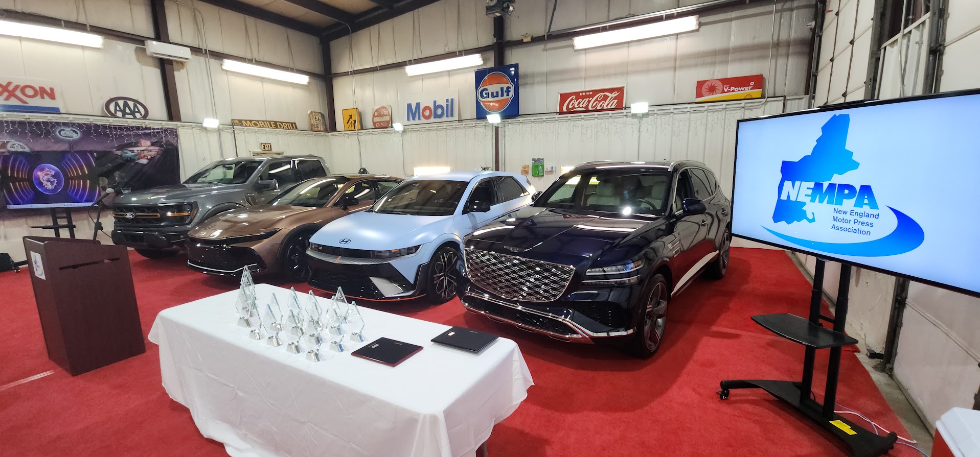 Four award-winning vehicles parked on a red carpet next to a table of glass trophies and a digital screen displaying the NEMPA logo.