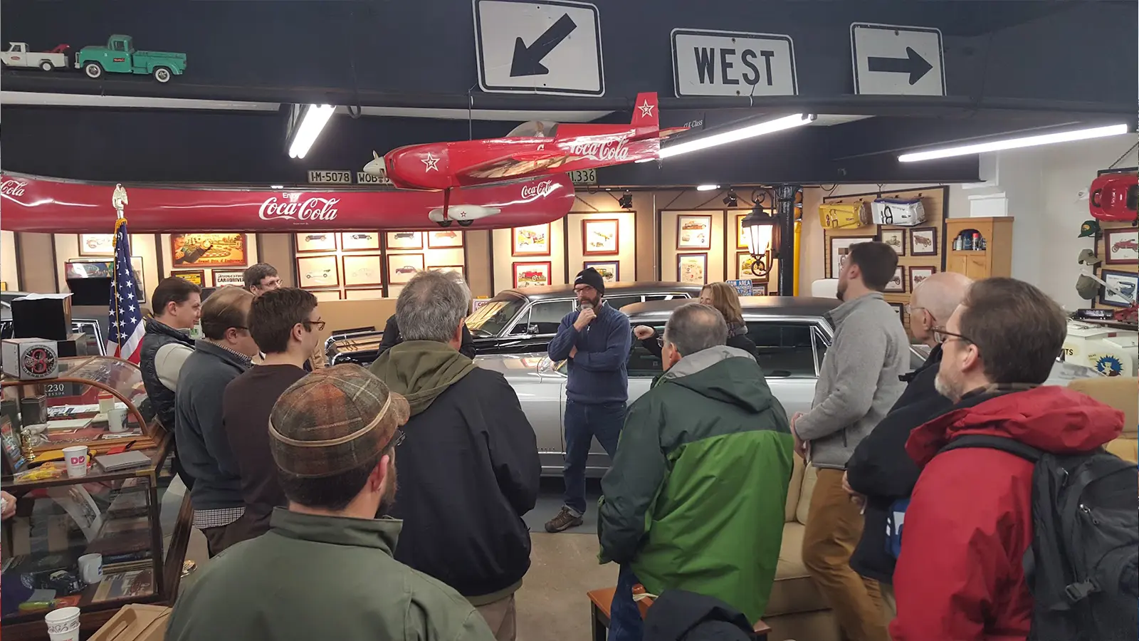 NEMPA members listen to a speaker beside a classic silver sedan inside the Automotion museum, beneath hanging Coca-Cola model airplanes and vintage road signs.